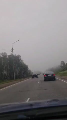 The video is shot from the perspective of someone inside a vehicle traveling on a foggy highway. The road ahead is visible with two cars in front, one black sedan and a smaller black vehicle. The left side of the road is lined with trees, and there's a lamppost with a circular element on it. The fog obscures the view of the horizon and the distance, creating a hazy atmosphere. The car the footage is filmed from is partially visible at the bottom of the frame, appearing to be the dashboard and windshield.