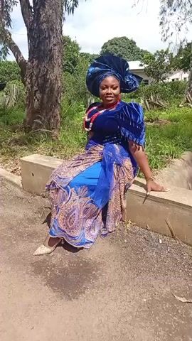 A woman sits on a concrete ledge outdoors, dressed in a blue and gold traditional outfit, complete with a large head wrap, jewelry, and matching shoes. She is leaning back slightly with her left arm resting on the ledge. The background shows a tree to her left, foliage and a building in the distance, and a dirt path in the foreground. The lighting suggests it is daytime with sunlight creating shadows.