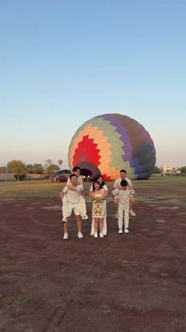 The video shows a group of six people standing in a field with a hot air balloon in the background. Four of the people are giving piggyback rides to the other two. Everyone is wearing white or light-colored clothing. The hot air balloon is multi-colored and partially inflated. The field is dirt and grass. There is a red car and some trees in the background. The sky is clear and blue.