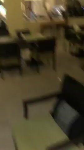 The camera pans around a well-lit dining area, showing a few dark-colored tables with matching chairs. Each table is set with placemats and napkins in a light green shade. The floor is polished and reflective, adding to the overall brightness.  In the background, there's a counter where a staff member appears to be working, and an open buffet area is visible further back.  The ceiling is dark with exposed beams, creating a contrast with the light walls and floors.