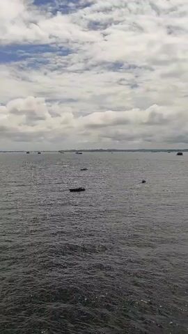 A panoramic video captures a wide expanse of choppy, dark water under a cloudy sky. Several small boats are scattered across the water, appearing as dark silhouettes. In the distance, a larger red and white cargo ship is visible, as well as a landmass with buildings and trees. The clouds are a mix of white and gray, suggesting a partly cloudy or overcast day. The video pans slowly to the right, revealing more of the water and distant landscape.