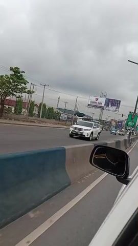The video shows a view from inside a car driving on a road. There are other vehicles on the road, including vans and cars. There are several billboards visible, advertising services like "USSD Lock," "Charles Dale International School," and "OPay Online Subscription Control." The sky is overcast.  A concrete barrier runs along the side of the road. The video appears to be shot in an urban area in Nigeria.