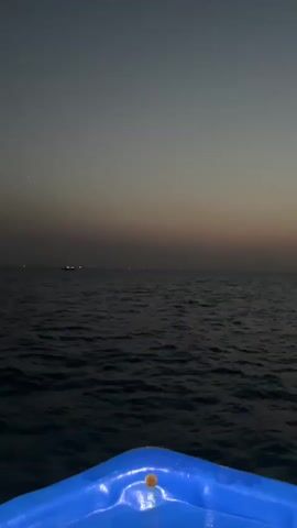 A first-person perspective video shows the bow of a blue boat moving across dark, choppy ocean water. The sky is a gradient of grey and faint orange, suggesting either dusk or dawn. In the distance, on the horizon, a few faint lights are visible, possibly indicating land or other vessels. The camera angle is tilted slightly downwards, with the majority of the frame dedicated to the sea and sky. The overall atmosphere is calm and serene.
