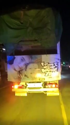 The video shows the back of a truck at night. The truck has a green tarp covering its cargo and two images on the back. The left image is of two children, one older and one younger, with the older child embracing the younger child. The right image is of a cartoon character with text written in a stylized font. The truck is moving on a road, and the headlights of other vehicles can be seen in the distance. The road appears to be wet, and there is a yellow glow from streetlights or other illumination. The overall scene is dark and somewhat blurry due to the movement and low light conditions.