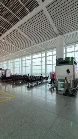 The video shows the interior of an airport departure lounge. Large windows look out onto an airfield where planes are visible. Rows of empty seats are arranged in the lounge, with a few passengers scattered throughout. One person is looking at their phone, another is walking with someone. An escalator is visible in the foreground. The lighting is bright and even, suggesting an overcast day. There is a large advertising display to the right and a smaller display showing flight information to the left.