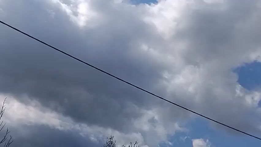 The video shows a shot of the sky with clouds, a utility pole with a street lamp, and some bare tree branches. The clouds are a mix of white and dark grey, indicating varied weather conditions. A single power line stretches across the frame. The street lamp is simple in design. The camera angle is low, looking upwards towards the sky.