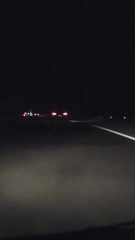 The video is a first-person view of a car driving on a highway at night. The headlights of the car illuminate the road ahead, revealing white lane markings. In the distance, other vehicles can be seen with their red taillights. The sky is dark and featureless. The overall atmosphere is dark and somewhat eerie.