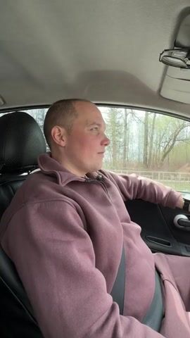 The video captures a man inside a moving car, appearing to be in the driver's seat, though he is filmed from the passenger side. He is wearing a mauve-colored tracksuit, consisting of a quarter-zip sweatshirt and matching bottoms, and is securely buckled into a black seatbelt. The man has a short-cropped hairstyle and a clean-shaven face.

Initially, he looks out the car window with a gentle smile, observing the passing scenery. He then turns to the camera, flashing a wide, joyful grin, before returning his gaze forward and out the window, maintaining an amused expression. The car's interior is dark, likely black or dark grey, with a textured headrest visible behind him.

Outside the car, through the windows, a daytime scene unfolds. Trees with mostly bare branches and some nascent green foliage line what appears to be a road or path, indicating spring or early summer. Another car can be seen passing in the opposite direction, and a light-colored fence is visible further back.

The audio features upbeat music with a clear Russian voice singing "С днём рождения" (S dnyom rozhdeniya), which translates to "Happy Birthday." The man's happy demeanor aligns with the celebratory nature of the song. The setting is identified as Rogachovo, Russia.