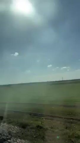The video shows a flat green field under a bright, slightly overcast sky. The sun shines intensely, creating a glare. Power lines are visible in the distance. The foreground shows some rough terrain or roadside.