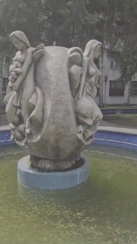 A statue of four women is in the center of a pond. The statue is made of stone and is worn, with a greenish tint. The women are all nude and are depicted in various poses, some appearing to be holding or interacting with other figures. The pond water is murky and green. In the background, there is a building with windows and trees. The lighting is dim, suggesting an overcast day.