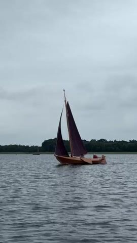 A wooden sailboat with a dark red sail is moving across a body of water under a cloudy sky. The boat has a single mast and two sails. There appear to be two people on board. In the distance, there are trees lining the shore and other sailboats. The water is choppy.