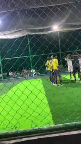 A soccer game appears to be on an artificial turf field.  A small crowd is present, though the view is partially obstructed by a green netting fence. Two teams face each other, huddled for pre-game interaction. One team is wearing yellow jerseys and the other is in white. A referee is present in black and white. The lighting is bright and even.