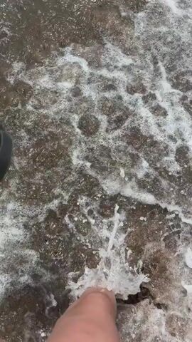 The video captures a first-person perspective at a sandy beach in Pylos, Greece, showcasing the shallow ocean water.

A person's bare leg and foot, with red nail polish, are seen wading in the foamy, incoming waves at the water's edge. The sand appears dark and wet. Around the 0:01 mark, the individual picks up a black, puffy sandal with "Ugg" branding clearly visible on its straps. The person continues to stand or walk in the shallow water, interacting with the gentle surf.

The final shot focuses on the waves breaking and receding onto the dark sand, highlighting the white foam against the brownish water. The daytime lighting suggests pleasant weather for beach activities, with the person enjoying the sea.