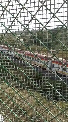 The video shows a train moving through a landscape, as seen through a chain-link fence. The train cars are primarily white with red and orange stripes. The landscape features greenery and hills. The fence is green and obscures the view slightly.