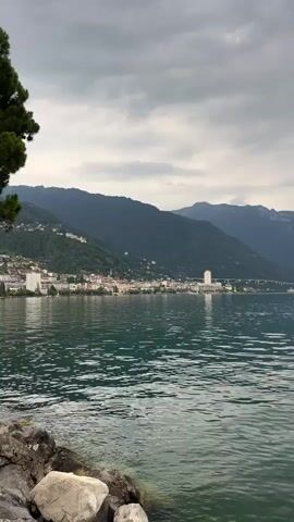 A slow pan across a wide, greenish lake with a rocky shoreline in the foreground. In the middle ground, a long, built-up town or city stretches along the base of steep, forested mountains. The sky is overcast with dark, heavy clouds. The far distance shows more hazy, layered mountains.