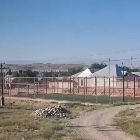 The video shows a fenced-in outdoor sports court with artificial turf. In the background, there are houses, trees, and hills under a clear blue sky. A single utility pole stands to the left of the court, and a pile of rocks is visible in the foreground. The camera pans from left to right, offering a view of the court and its surroundings.