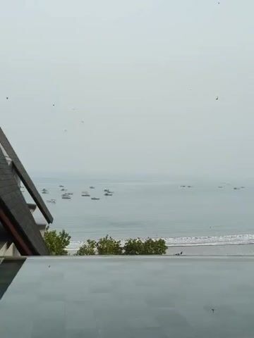The video shows a panoramic view of the ocean from a high vantage point, likely a balcony or rooftop. In the foreground, a dark, tiled surface, possibly a swimming pool edge, is visible. The sea is calm, with small waves breaking on the shore. Numerous small boats are scattered across the water, suggesting they are fishing boats or small vessels. A stretch of sandy beach is visible beyond the waves, with a few scattered trees and some beach umbrellas. The sky is overcast and hazy, giving the scene a muted, serene atmosphere.
