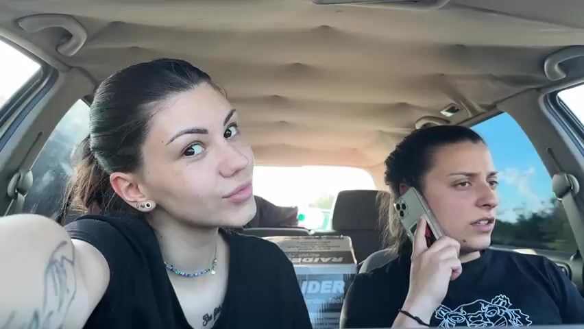 Two young women are sitting in the front seats of a car. The car appears to be stationary. The woman on the left is looking at the camera and gesturing. She is wearing a black t-shirt and a blue beaded necklace. Her hair is pulled back in a ponytail. The woman on the right is talking on a cell phone. She also has her hair in a ponytail and is wearing a black t-shirt. In the back seat are boxes of what appears to be gardening tools. The interior of the car appears worn. The sky is visible through the windshield.