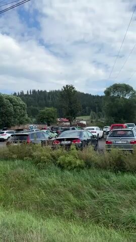 The video pans across a parking lot filled with numerous cars, suggesting a populated area or an event. In the background, a sign with the word "ZOO" is visible, indicating the presence of a zoo. Beyond the parking lot and the zoo entrance, a lush green forest and hills can be seen, creating a natural and scenic backdrop. The sky is partially cloudy, with patches of blue visible. The overall impression is a bustling day at a zoo, surrounded by nature.