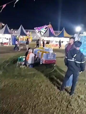 The video shows an outdoor night scene with several people milling around. There are carnival-style rides, including a small train and a carousel. Tents and booths are also visible, with some decorated with lights. The ground appears to be grassy. There are colorful pennant flags strung up in the air. The overall atmosphere suggests a fair or carnival at night.