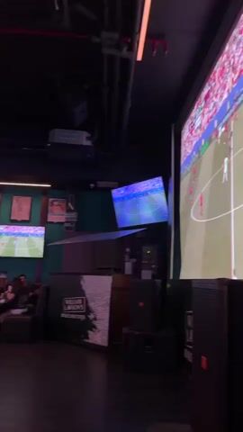 The video displays a lively indoor establishment, possibly a sports bar or restaurant, filled with patrons. The environment is dimly lit, with neon strip lights illuminating the ceiling and the top edge of the walls. Multiple flat-screen televisions are mounted on the walls, displaying sports games, specifically football matches. Flags of various countries hang from the ceiling and walls, suggesting an international sporting event is being broadcasted or celebrated. Several tables with chairs are occupied by people who are seated, interacting, and watching the screens. Some individuals are seen holding drinks and appear engaged in conversation. The decor includes some framed artwork or posters on the walls. The overall atmosphere suggests a social gathering where people are enjoying entertainment and refreshments.
