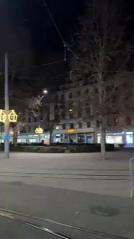 The video shows a street at night in what appears to be a European city. Several multi-story buildings line the street, with lit-up signs indicating businesses. There are street lamps with decorative lighting, and bare trees are visible. The street has tram lines running along it. In the latter part of the video, a group of people wearing what look like police or security uniforms are standing on the street. The scene is generally quiet, with a few cars passing in the background.