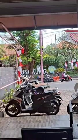 The video shows a street scene viewed from a covered area. In the foreground, several parked motorcycles are visible, including a white and red one and a black one with a black helmet on top. To the left, a metal fence with red and white striped flags is present. Beyond the motorcycles, a street is visible with a white SUV driving away and a turquoise truck with a black cab passing by. Several people on motorcycles are also seen on the street. In the background, there are trees and a building with a sign that reads "KUNG". Red and white flags are also displayed on poles in the background. The overall environment appears to be a public street in what may be a Southeast Asian country, given the presence of the motorcycles and the red and white flags, which are reminiscent of the Indonesian flag.