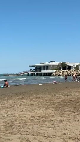 A wide shot shows a sandy beach on a clear, sunny day. The ocean is visible with moderate waves. In the distance, a modern, white building with large windows stands on stilts over the water, accessible by a wooden walkway. Several people are on the beach, including a person sitting alone on the sand in the foreground, and a group of people, including children, playing near the water's edge. Beach chairs and towels are also visible. The sky is a clear, bright blue.