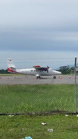 A small propeller plane, white with red accents, sits on an airfield tarmac behind a chain-link fence. Orange traffic cones are placed on the tarmac around the plane. In the background, beyond the tarmac, are several industrial-looking buildings and a control tower. A windsock, orange and white, flies from a pole in front of the control tower. The sky is overcast. Green grass and some scattered litter are visible in the foreground. The camera pans slowly from left to right.