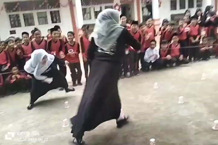 The video shows an outdoor school event with a group of children and adults gathered. In the foreground, two women in black hijabs and long dresses are participating in a game, navigating around small white cups placed on the ground. The woman on the left is in motion, running and looking towards the right. The woman on the right is also in motion, facing towards the left and moving away from the camera. Behind them, a line of children, mostly boys in red shirts and black pants, and girls in black hijabs and dresses, are seated or standing, watching the game. Some adults, also wearing hijabs, are also present among the spectators. The environment appears to be a school courtyard, with a building in the background decorated with red and white banners. The ground is paved, and there are some trees and plants visible. The overall atmosphere suggests a festive or celebratory occasion.