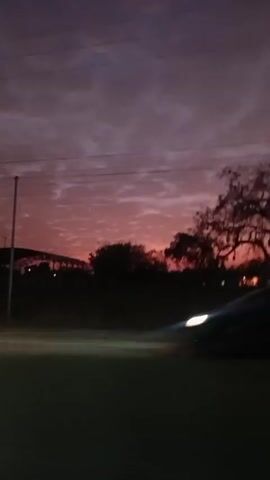 The video shows a dark landscape during dusk or dawn. The sky is a blend of dark purple and fading pink colors with clouds covering most of the visible area. Silhouettes of trees and sparse structures are visible against the colorful sky. There are a few blurry lights in the distance, possibly from buildings or streetlights. A dark, indistinct shape of a car can be seen briefly at the beginning of the video. The camera appears to be moving horizontally, possibly from inside a vehicle.