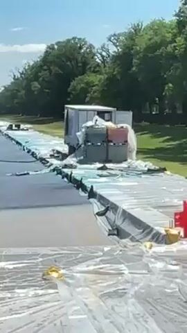 The video captures an outdoor scene on a clear, sunny day, likely on the National Mall in Washington D.C., with the Washington Monument and the distant United States Capitol building visible in the background. A large expanse of ground is undergoing construction or preparation, with sections covered in white plastic sheeting. A dark-colored pickup truck, towing a trailer equipped with a large tank, drives from left to right across a freshly paved or painted surface that displays shades of dark blue and lighter blue. In the mid-ground, several white utility trailers or containers are stationed, and further down the path, other utility vehicles and a few workers are visible. Orange traffic cones mark specific areas on the ground. The sky is bright blue with a few scattered white clouds.