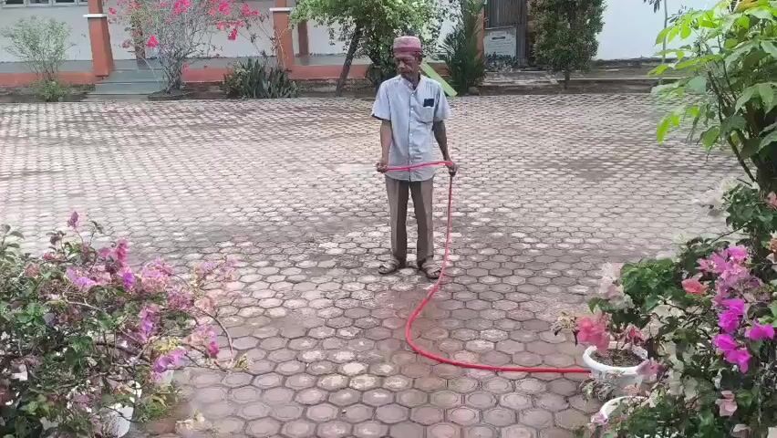 A man in a light blue shirt, light brown pants, and a maroon cap stands in a paved courtyard, holding a red hose. The hose snakes across the ground towards the right side of the frame. The man appears to be watering the plants in large white pots, which are visible in the foreground on both the left and right sides of the frame. The plants have abundant pink and purple flowers. In the background, there is a building with a beige facade and a few trees. The ground is wet, indicating recent watering.