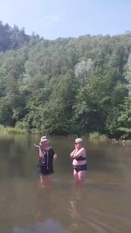 Two women stand in shallow water. The woman on the left wears a black dress and a white hat. The woman on the right wears a black bikini. Behind them, a wooded hill rises steeply. The sky is clear. The water is murky.