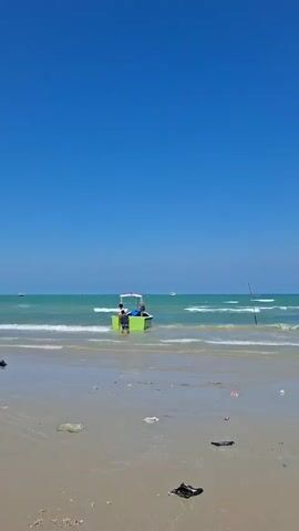 The video shows a beach with a bright blue sky. There are waves coming in from the ocean onto a sandy shore. The sand is littered with various pieces of trash, including plastic bags and wrappers. In the water, near the shore, there is a small, bright green boat. Two people, wearing t-shirts and shorts, are standing in or next to the boat, appearing to interact with it. One person is wearing a white shirt and blue shorts, and the other is wearing a darker shirt and shorts. A fishing rod is visible on the beach to the right of the boat. In the distance, there are more boats and what appears to be a coastline with buildings. The water is a clear turquoise color.