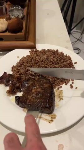 The video shows a white plate filled with a steak, a pile of buckwheat, and a scattering of what appears to be fried onions or bacon. A hand is holding a fork to stabilize the steak while another hand uses an IKEA knife to slice into it. In the background, a wooden tray holds several small containers and two bread rolls, while a power cord is visible on the floor. The table beneath the plate is white and has a few small brown spots on it.