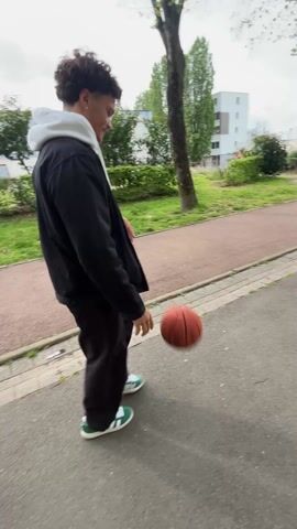 A young man walks along a sidewalk in a park, dribbling a basketball. He has curly hair and is wearing a white hoodie, a black jacket, black pants, and green and white sneakers. The sidewalk is next to a grassy area with trees, and a red pedestrian path runs parallel to the sidewalk. In the background are apartment buildings. The sky is overcast.