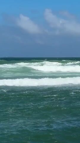 The video displays a wide shot of the ocean on a bright, partially cloudy day. The sea exhibits a deep green to turquoise hue, with numerous waves rolling in from the distance and breaking into white foam closer to the foreground. The waves vary in size, with some cresting significantly. The sky above is a clear blue, dotted with scattered white clouds. No land, people, or objects are visible in the frame, emphasizing the vastness and dynamic movement of the ocean. The scene is calm but with noticeable wave action, suggesting a gentle breeze or moderate sea conditions.
