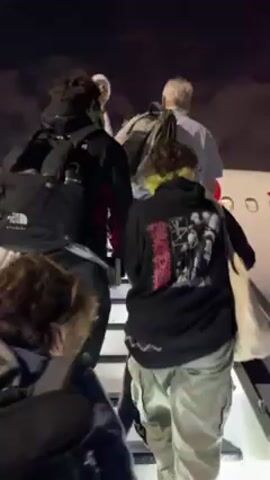 The video shows passengers boarding an Avianca airplane at night. The first shot is from behind, looking up the stairs toward the entrance of the plane as passengers ascend. The next shot shows the side of the plane, including the Avianca logo, the engine, and the tail. The plane is white with red accents. There are orange cones near the plane, and a vehicle attached to the plane near the engine. The background shows a dark night sky.