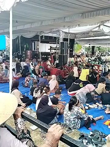 A large group of people, many wearing traditional clothing and head coverings, are gathered under a white tent. They are sitting on a blue tarp spread on the ground, watching a performance or event taking place on a stage in the background. The stage is set up with sound equipment, including large speakers, and there are performers on the stage. Some people in the foreground are wearing conical hats, suggesting a rural or outdoor setting. The overall atmosphere appears to be one of a community gathering or celebration.