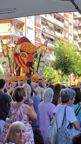 A flower float depicting a large, ornate buffalo with antlers is being displayed in a street parade. The buffalo is adorned with red, orange, yellow, and green flowers, and its eyes are large and red. Two smaller figures, possibly deer or reindeer, flank the buffalo. People are gathered on both sides of the street, many holding up phones to take pictures and videos of the float. The crowd is diverse in age and gender, and they appear to be watching the parade with interest. The buildings in the background are multistory and have balconies with people watching from above. The day appears to be sunny and warm, suggesting an outdoor event. The overall atmosphere seems festive.