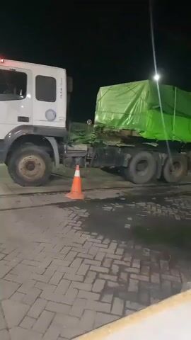 A semi-trailer truck is parked at night on a paved surface made of interlocking bricks. The truck's trailer is covered with a bright green tarp. An orange traffic cone is placed near the truck's rear wheels. The ground around the truck shows dark, wet patches, possibly from a spill. A bright light fixture is visible overhead, illuminating the scene. The truck has multiple axles and wheels, and the cab is white. The tarp on the trailer has a logo or text printed on it, which appears to be red and white.