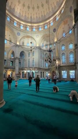 A wide interior shot shows the inside of a mosque with a large domed ceiling. The ceiling is ornately decorated with a large circular motif at the center and smaller repeating patterns around the dome. Several pendant lights hang from the ceiling. Arched windows are visible along the upper walls, and ornate detailing is present on the walls and arches. A vibrant teal carpet covers the floor, with rows of prayer rugs visible. Several people are present, engaged in prayer or walking. One person is bowing on the carpet in the foreground. Another person is kneeling. Several other individuals are walking across the carpeted area, and two children are among the people. The lighting appears to be natural light coming through the windows, supplemented by artificial light fixtures. The atmosphere suggests a place of worship.