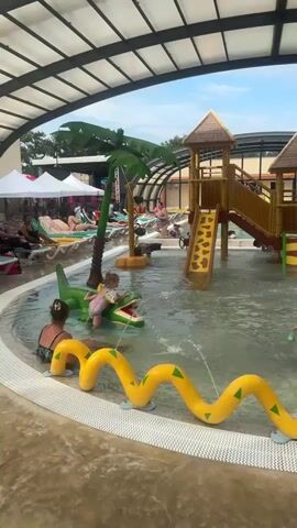 A children's splash pad is depicted, covered by a transparent, arched roof. In the center, a shallow pool contains several children playing. A young boy in swim trunks splashes in the water, while a toddler sits on a green alligator float, also in the water. Another child, wearing a pink swimsuit, is partially visible near the alligator. A woman with dark hair is seated at the edge of the pool, facing away from the camera. In the background, a large, multi-level wooden playground structure with slides is visible. To the right of the playground, a brown monkey sculpture is positioned near a waterfall feature. A yellow, wavy snake-like structure with green triangles runs along the edge of the pool in the foreground. Lounging chairs and people are visible in the background, suggesting a larger pool area beyond the splash pad. The overall environment appears to be a recreational water park or resort.