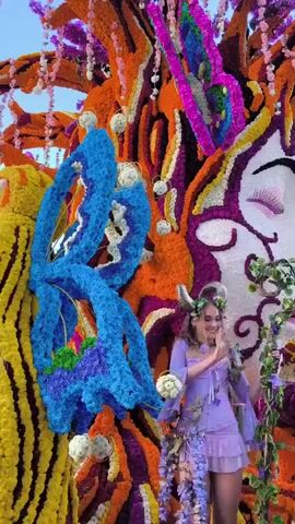 A woman in a purple fairy costume with wings stands in front of a large, elaborate floral display. The display features a giant face made of white and purple flowers, with closed eyes. Two large, blue butterflies made of flowers are positioned to the left of the face, and a smaller, pink butterfly made of flowers is at the top. The woman smiles as she holds a bouquet of white flowers. Vines and purple flowers hang from her costume. The floral display is vibrant and colorful, with oranges, purples, blues, and yellows. The lighting suggests it is daytime.