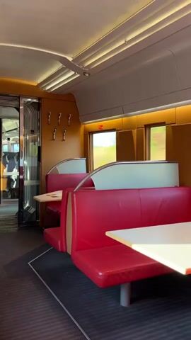 The video shows the interior of a train car, specifically the dining or lounge area. The seating consists of red leather booths with white table tops. The booths are arranged facing each other, with windows behind them that look out onto a blurry green landscape, indicating the train is in motion. The ceiling has a ribbed design with recessed lighting. There are coat hooks on a wooden panel. The overall aesthetic appears to be modern and comfortable. The footage is static, capturing a still scene.