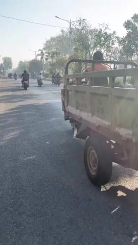 A three-wheeled utility vehicle drives down a paved road. The vehicle is olive green with a metal cargo bed and a black metal frame. A person wearing an orange shirt is visible in the driver's seat. The vehicle appears to be moving away from the camera. The road is lined with trees and there are other vehicles, including motorcycles, on the road in the background. The sky is clear and the lighting suggests it is daytime. The road appears to be in an urban or suburban environment.