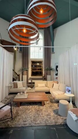 The video showcases a well-appointed living room setup within a spacious interior, likely a showroom or a repurposed historical building in Noordwijk, Netherlands. The scene is dominated by a large, light-colored L-shaped sectional sofa and a smaller matching sofa, positioned around a substantial wooden coffee table on a plush, light-colored shag rug. Two large, striking circular pendant lights with orange inner segments hang from a high, dark teal/green beamed ceiling, providing illumination.

At the back, a tall arched window is framed by long, neutral-toned curtains, allowing natural light to enter. Below the window, a console table features decorative items, possibly large mirrors, and vases with floral arrangements. Various small white side tables and light-colored ottomans are strategically placed. The room has white walls and a dark floor, possibly slate or stone. White fabric room dividers create a defined area for the living room display. No people are visible, and the video presents a static view of the designed space. A small, indistinct card with text is visible on a side table.