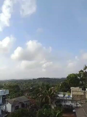 The video shows a wide outdoor shot of a landscape with buildings and trees under a cloudy sky. The camera is looking down on a few residential buildings in the foreground and a dense, green forest or jungle in the background. The sky is a light blue with scattered white clouds. The sun is visible behind some of the clouds, casting a bright light on the scene. The overall impression is of a tropical or subtropical environment.