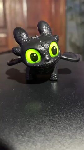 A close-up shot captures a black toy dragon with large, bright green eyes. The dragon is positioned in the center of the frame, facing forward, with its wings slightly extended. The background is blurred, with hints of wooden furniture and a dimly lit environment. The toy rests on a dark, textured surface. The video is short, looping the same image multiple times.